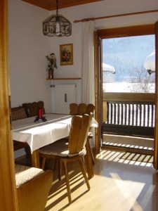 Blick in die Küche. Man sieht einen Tisch mit Eckbank und Stühlen und im Hintergrund den Balkon mit Blick auf eine winterliche Landschaft.